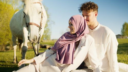 Free A loving couple sitting together in a field, smiling under a blue sky with a horse nearby. Stock Photo
