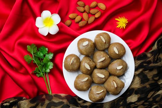 Plate of traditional sweets garnished with almonds, surrounded by flowers and nuts on vibrant red fabric.