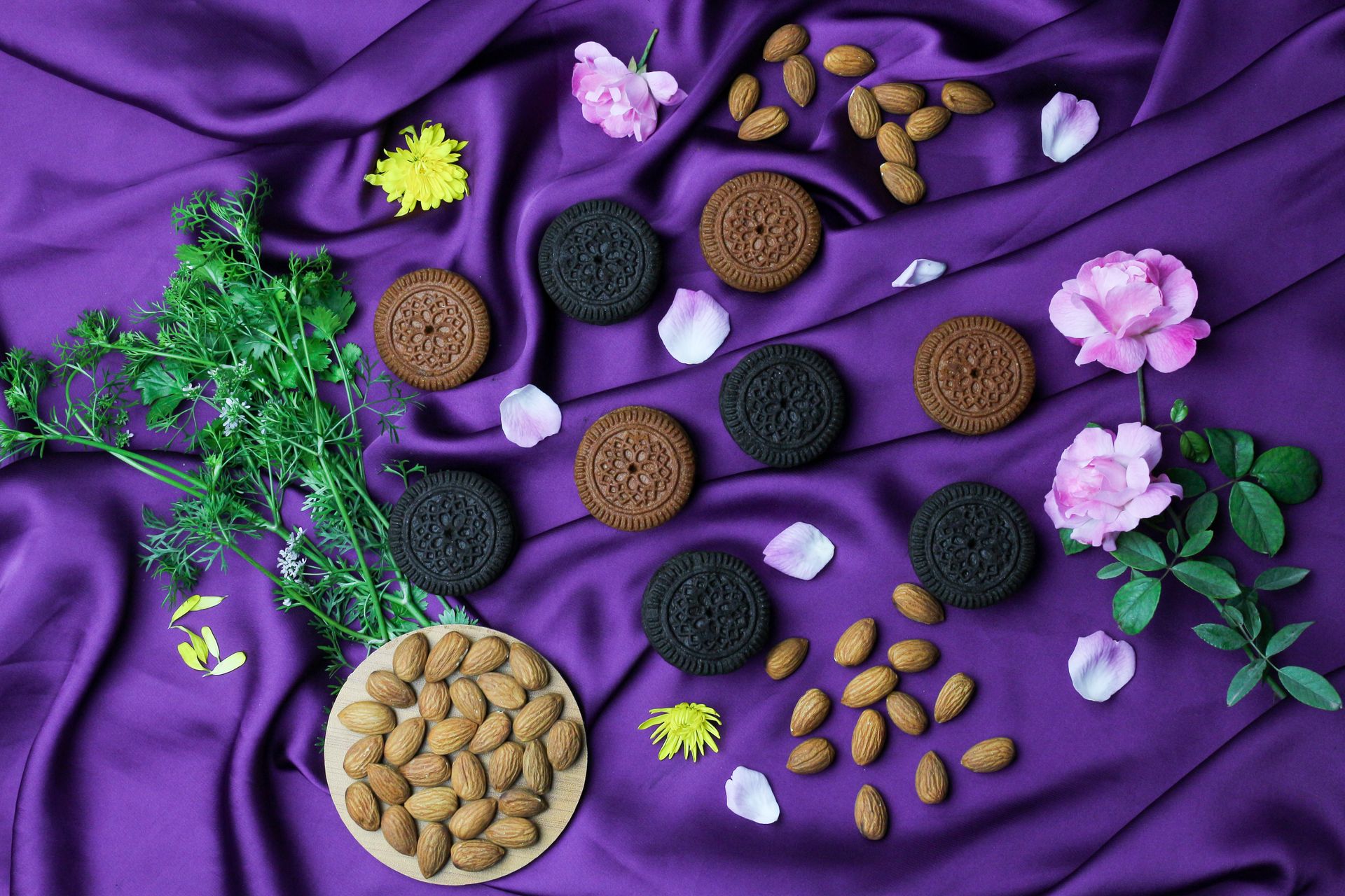 Artistic cookies and almonds arrangement on purple satin, with pink flowers and green herbs for a vibrant, elegant look.