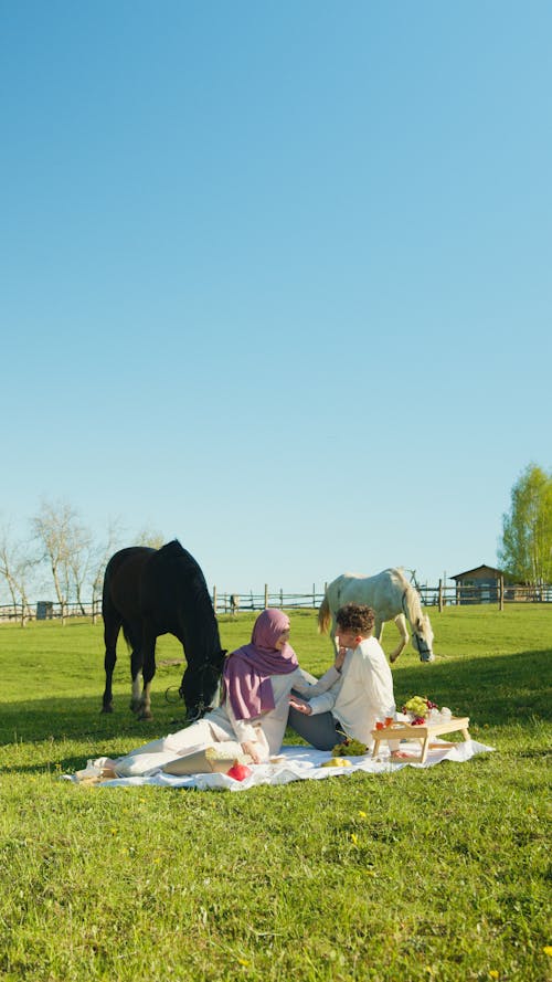 A Couple Sitting on  a Picnic Blanket Near Horses 