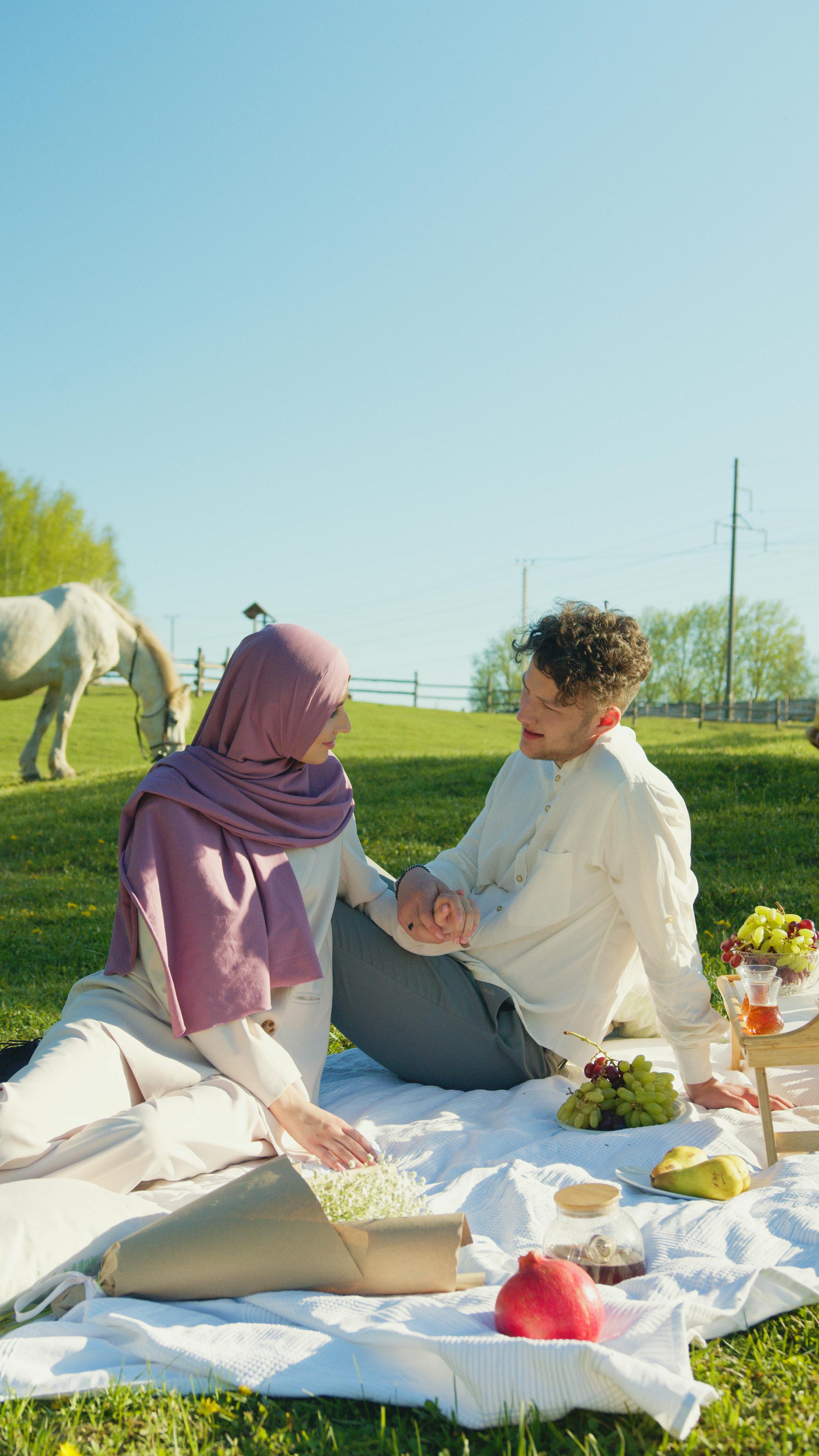 Couple having a romantic picnic in a sunny field, surrounded by nature and a grazing horse.
