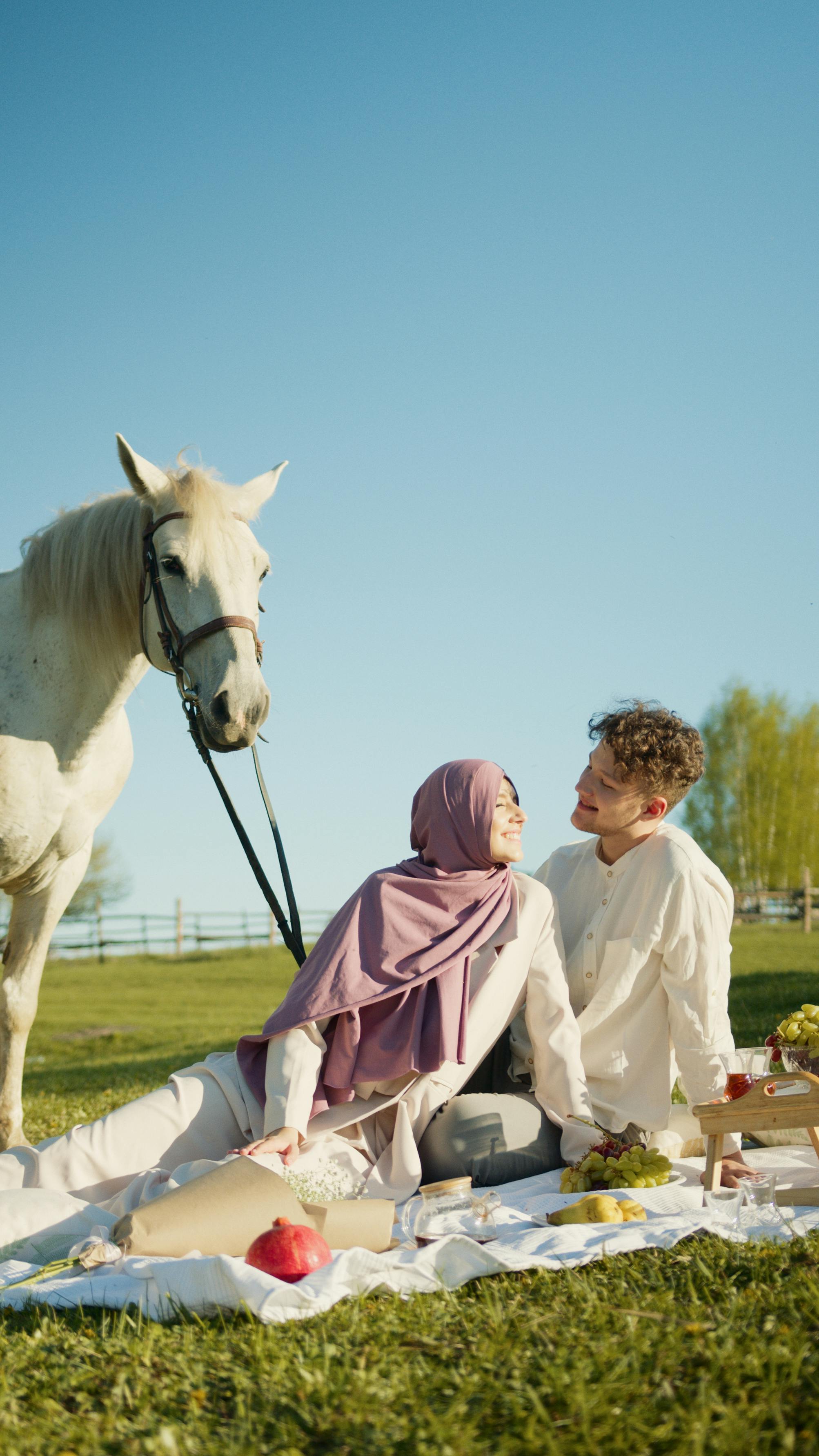 A couple enjoys a picnic with a white horse in a sunny green field.