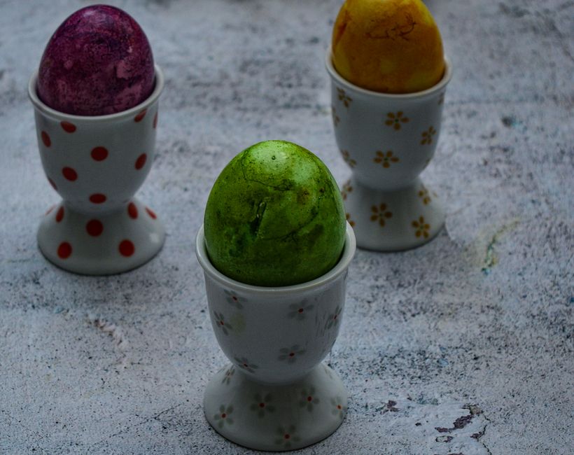 A dozen hard-boiled eggs in shades of pink, blue, yellow, and green, drying on a wire rack next to cups of colored dye water.