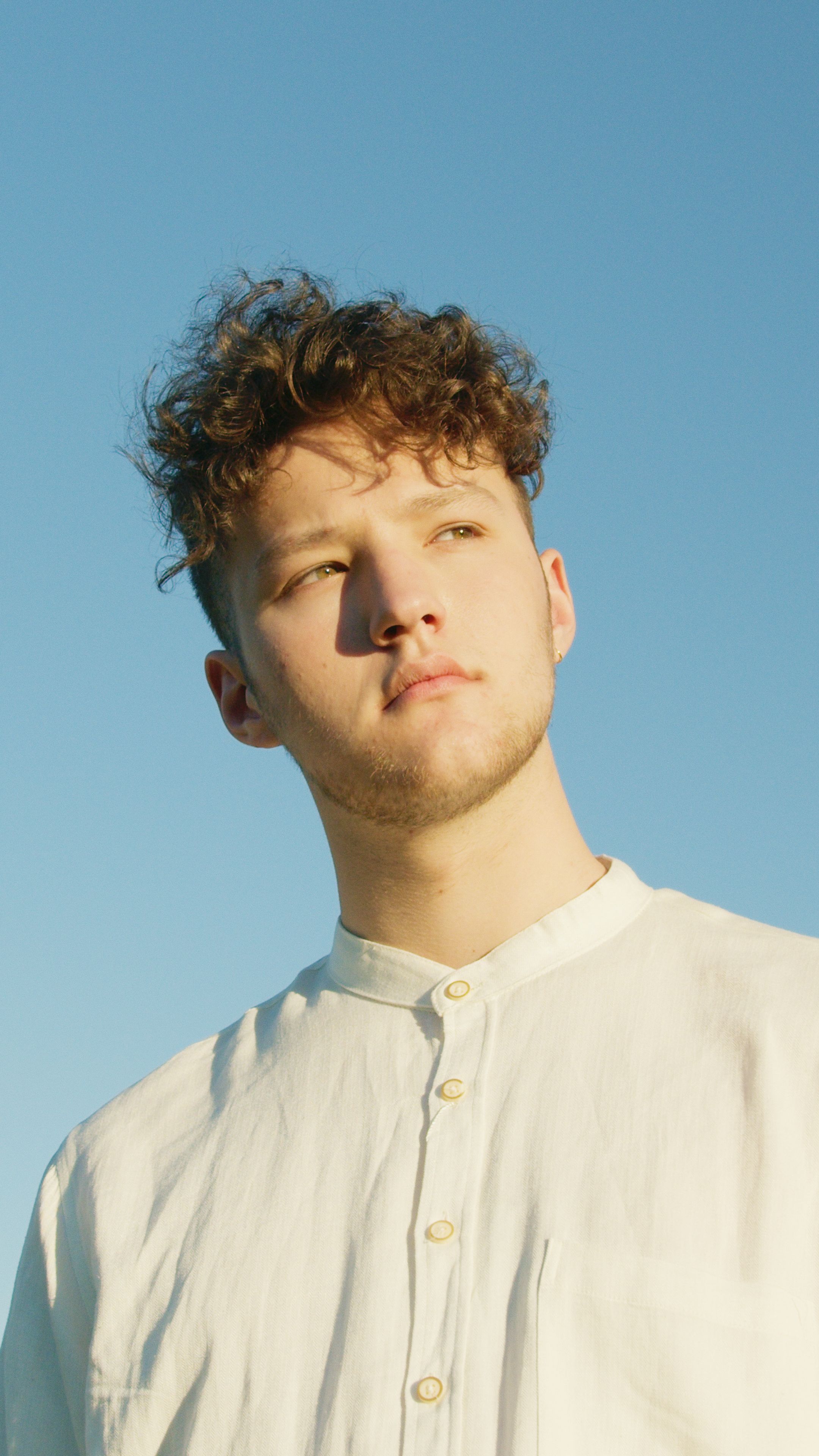 Portrait of a young man with curly hair gazing upwards, wearing a white shirt outdoors.