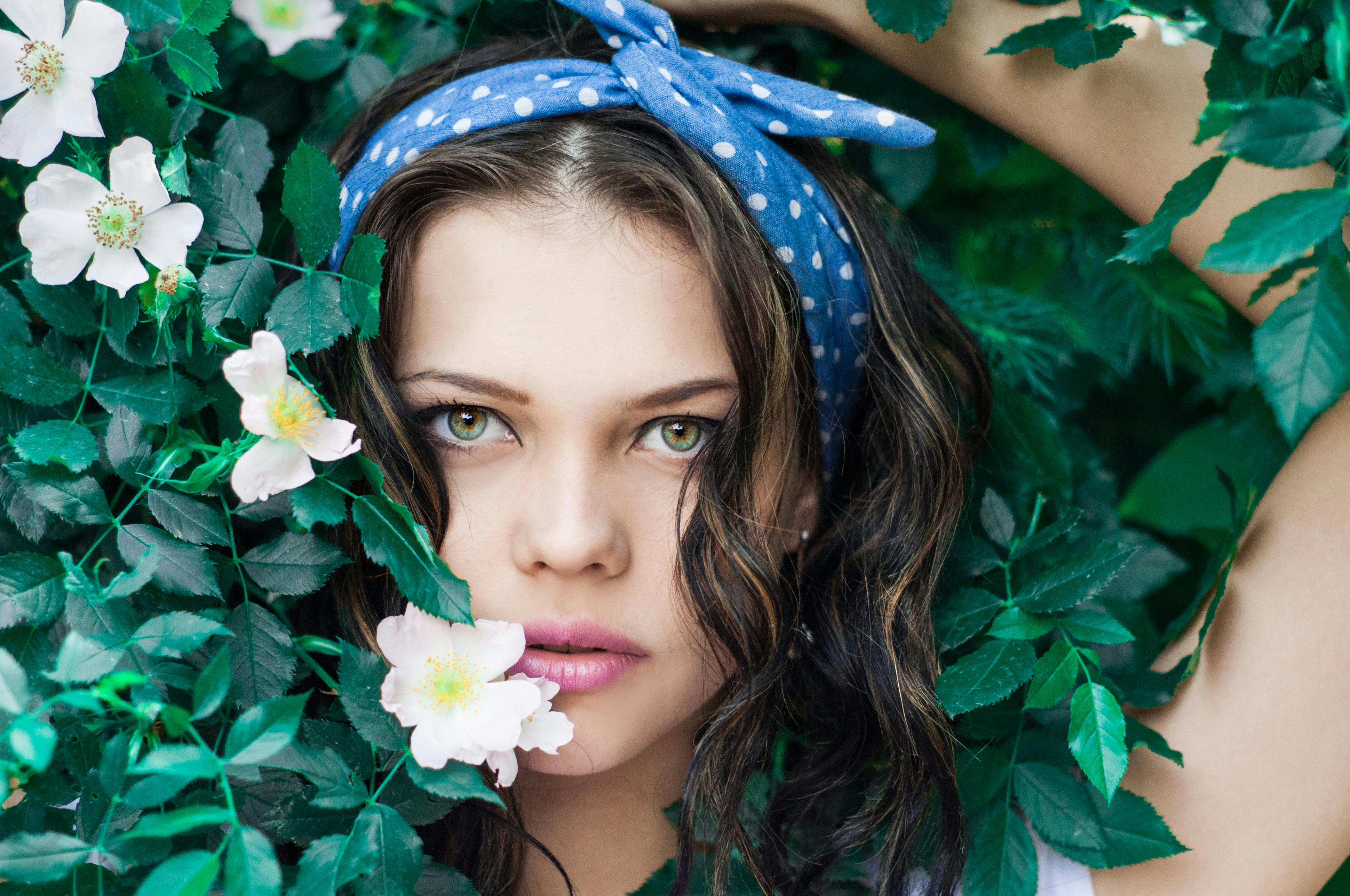 Young woman with polka dot headband surrounded by lush green leaves and white flowers.