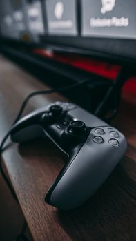 Close-up of a modern gaming controller on a wooden desk with blurred TV in background.