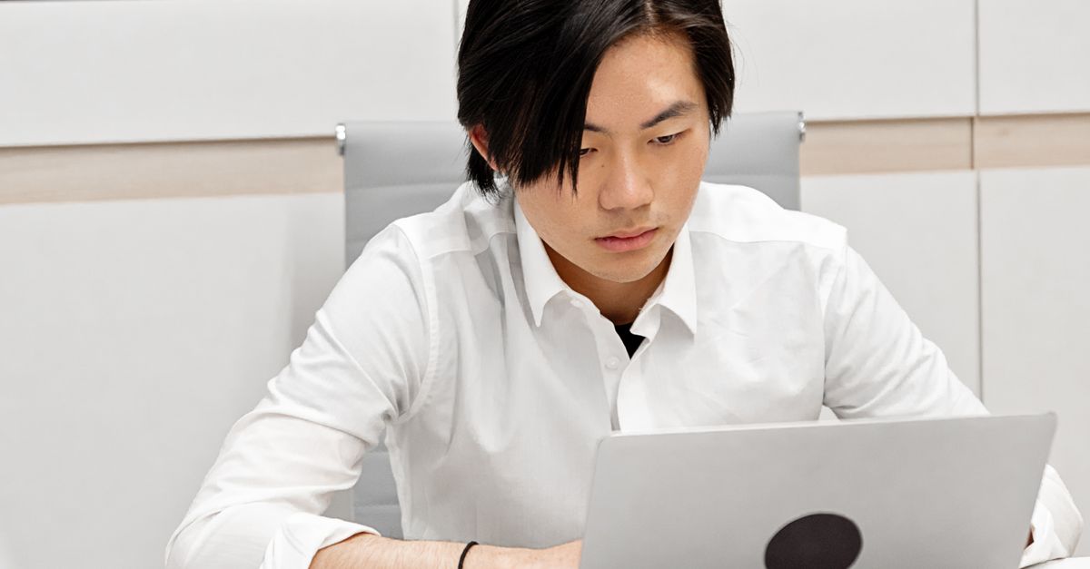 Asian man in white shirt working on laptop in modern office setting, focused and engaged.