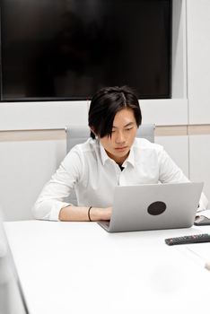 Asian man in white shirt working on laptop in modern office setting, focused and engaged.