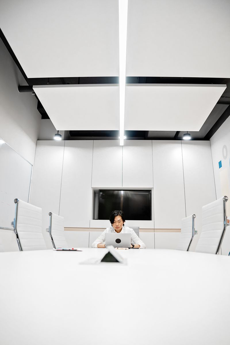 Asian man working on a laptop in a sleek conference room setting, focused on task.
