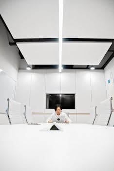 Asian man working on a laptop in a sleek conference room setting, focused on task.