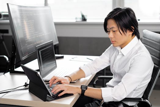 Asian man in white shirt focused on coding at modern office workspace.