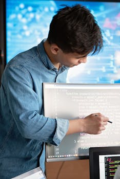 Asian male programmer writing code on a computer monitor in an office setting.