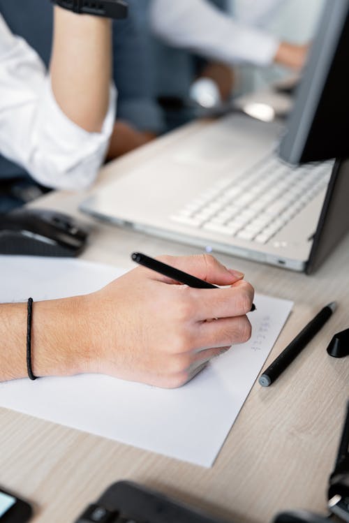 A Person Writing on a Bond Paper Beside a Laptop