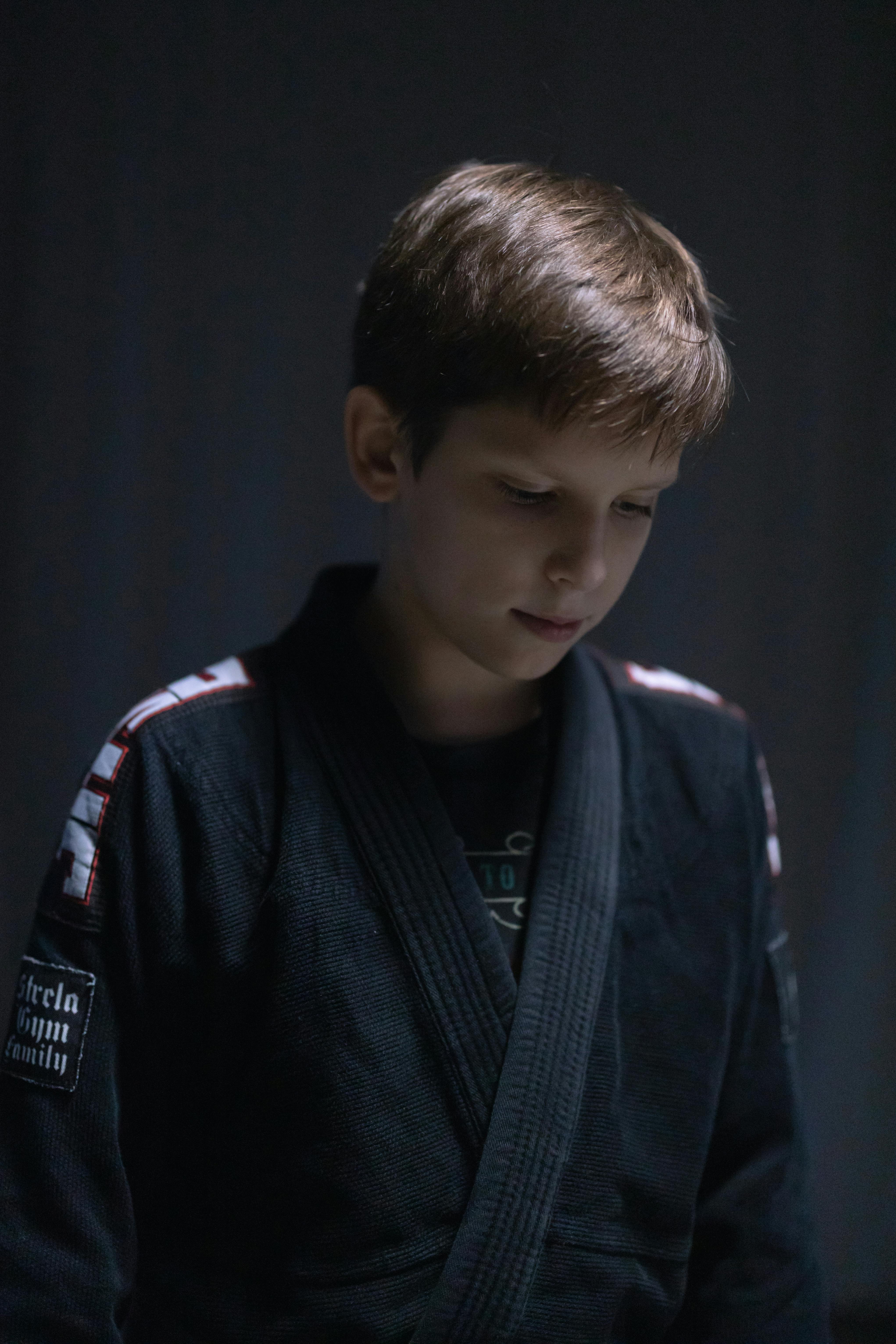 A contemplative young boy in a black jiu jitsu gi looks down with focus.