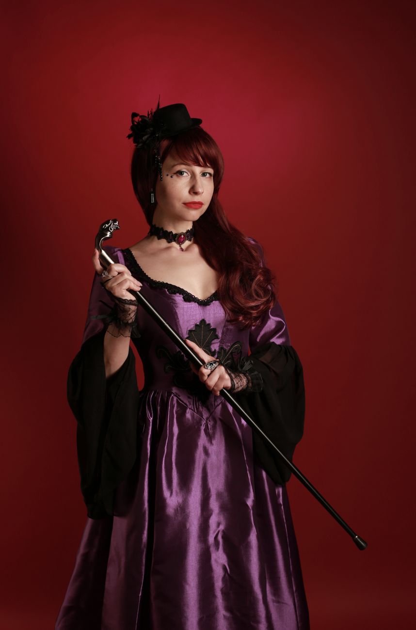 Woman in a purple Victorian-style gown holds a cane, set against a red background.. woman, purple, victorian