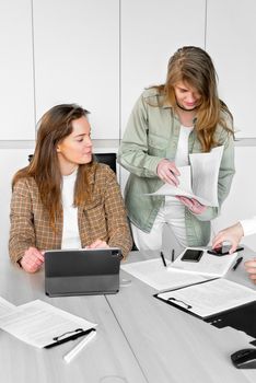 Two women discuss paperwork during a business meeting in a modern office setting.
