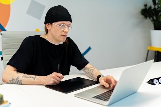 Tattooed designer working on a laptop and tablet in a stylish office setting.