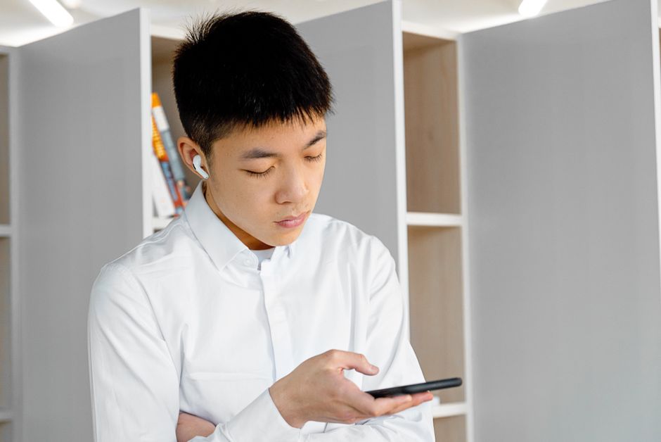 An Asian man in a white shirt using a smartphone with earbuds in an office setting.