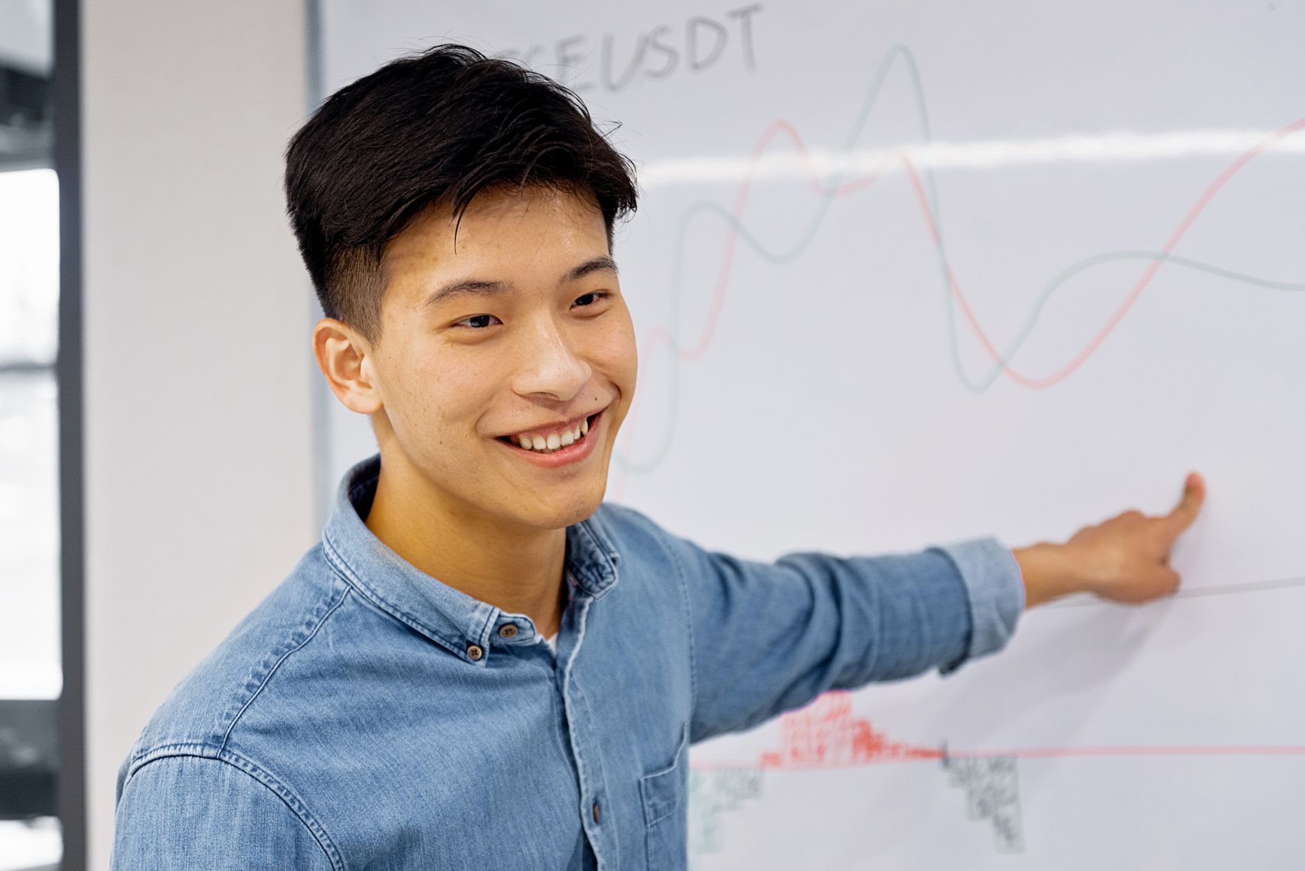 Smiling young Asian man presenting data on a whiteboard in a modern office setting.