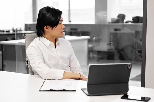 Free Asian businessman sitting in modern office, working on a tablet. Side view focused on the workplace environment. Stock Photo