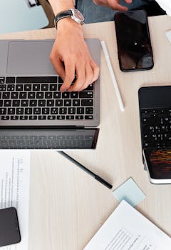High-angle view of a person typing on a laptop in an office setting with gadgets and documents.