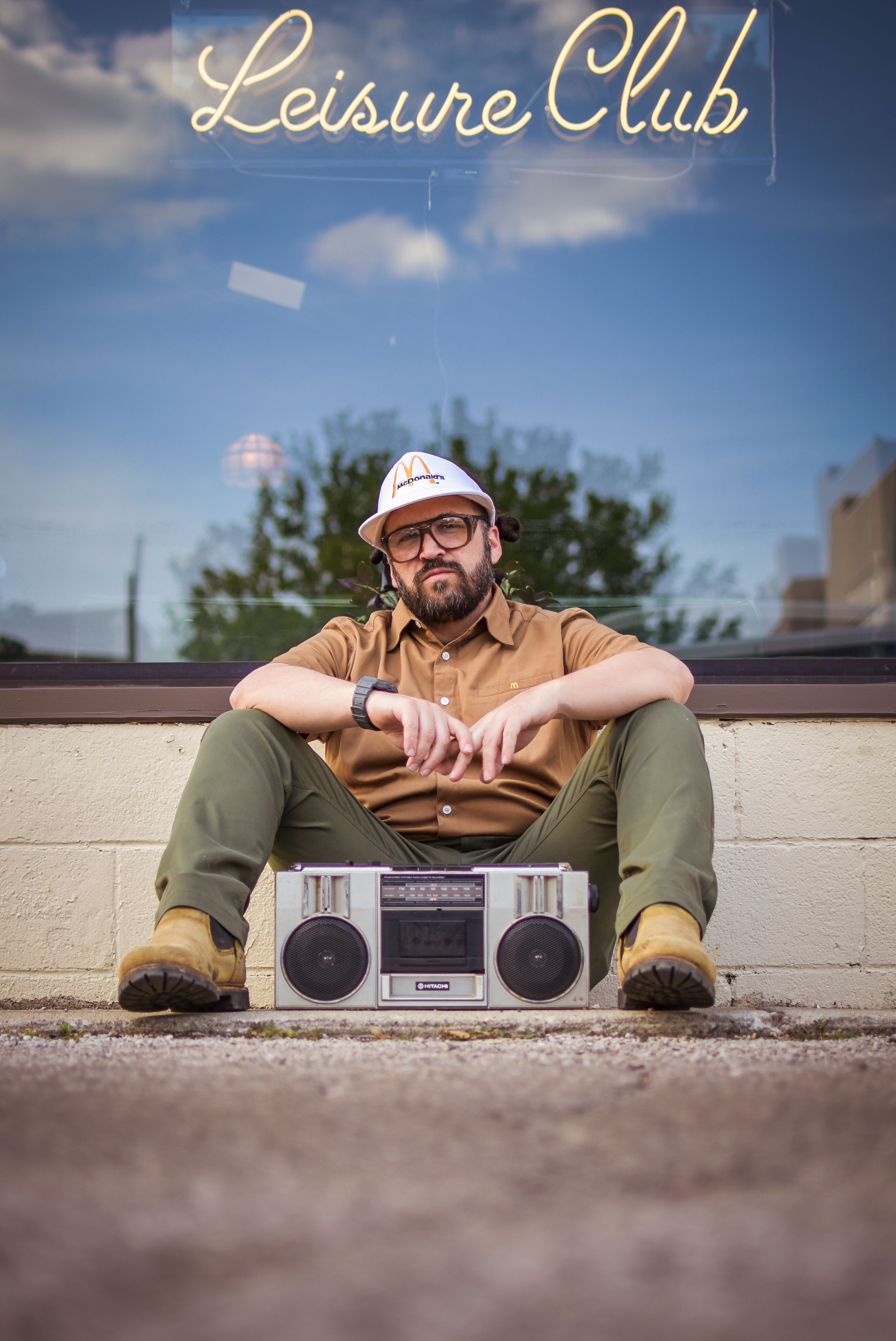 Bearded man sitting with boombox by glass window, wearing a cap at Leisure Club.