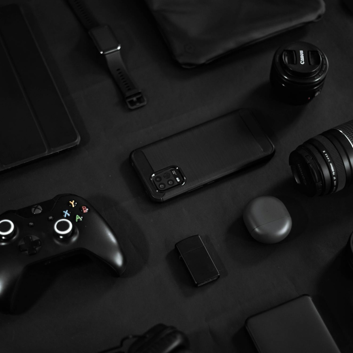Stylish arrangement of gadgets including a smartphone, lens, and game controller on a black surface.