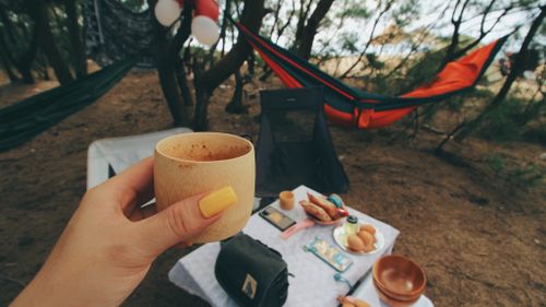 Free Cozy camping setup with a hand holding a wooden cup, hammocks, and picnic items under trees. Stock Photo
