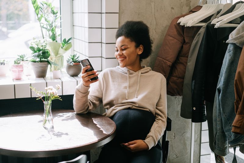 A Black pregnant woman with afro hair sits at a small table near a sunlit window, smiling at her smartphone with one hand resting on her belly.