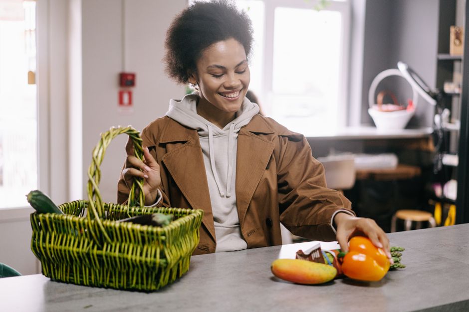 A woman joyfully choosing fruits and vegetables while grocery shopping indoors with a green basket.
