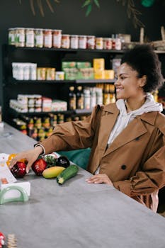 Woman with afro hair in a brown coat selecting fresh vegetables at a store counter, smiling.