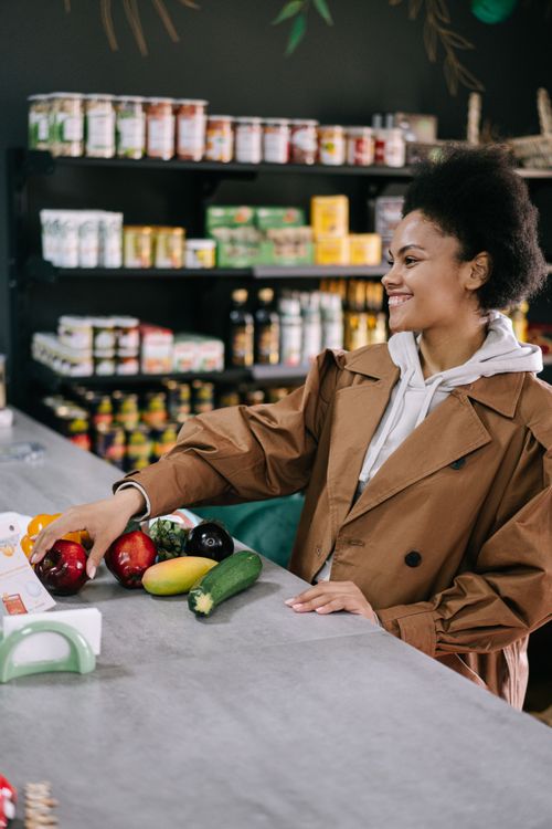Free Woman with afro hair in a brown coat selecting fresh vegetables at a store counter, smiling. Stock Photo