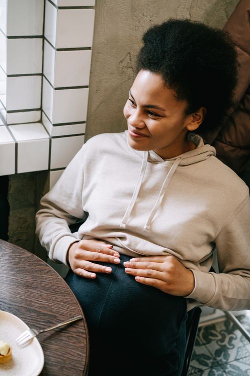 Free A serene pregnant woman with afro hair smiling and sitting by a table indoors. Stock Photo