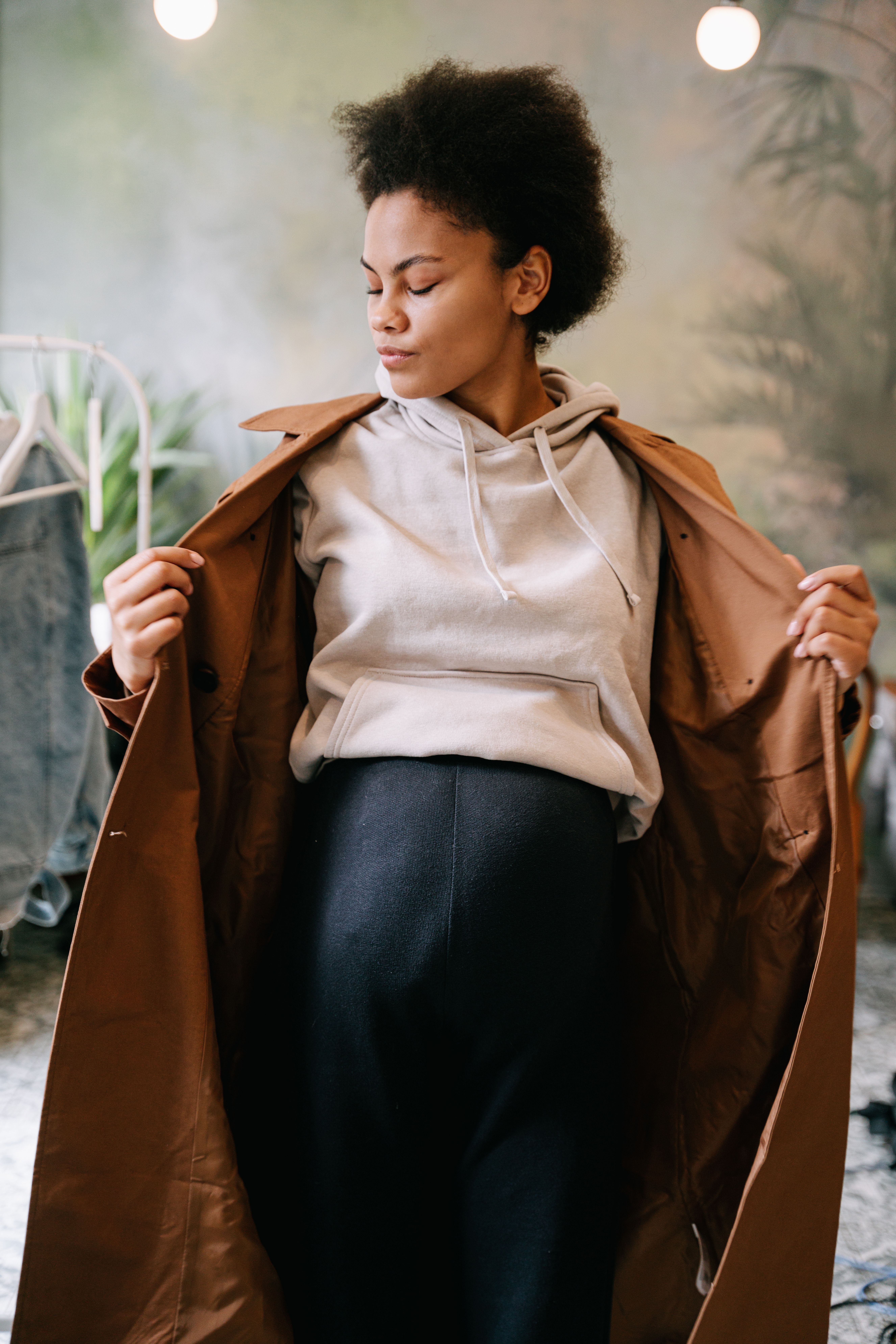 A pregnant woman adjusts her coat over a hoodie in a cozy indoor setting.