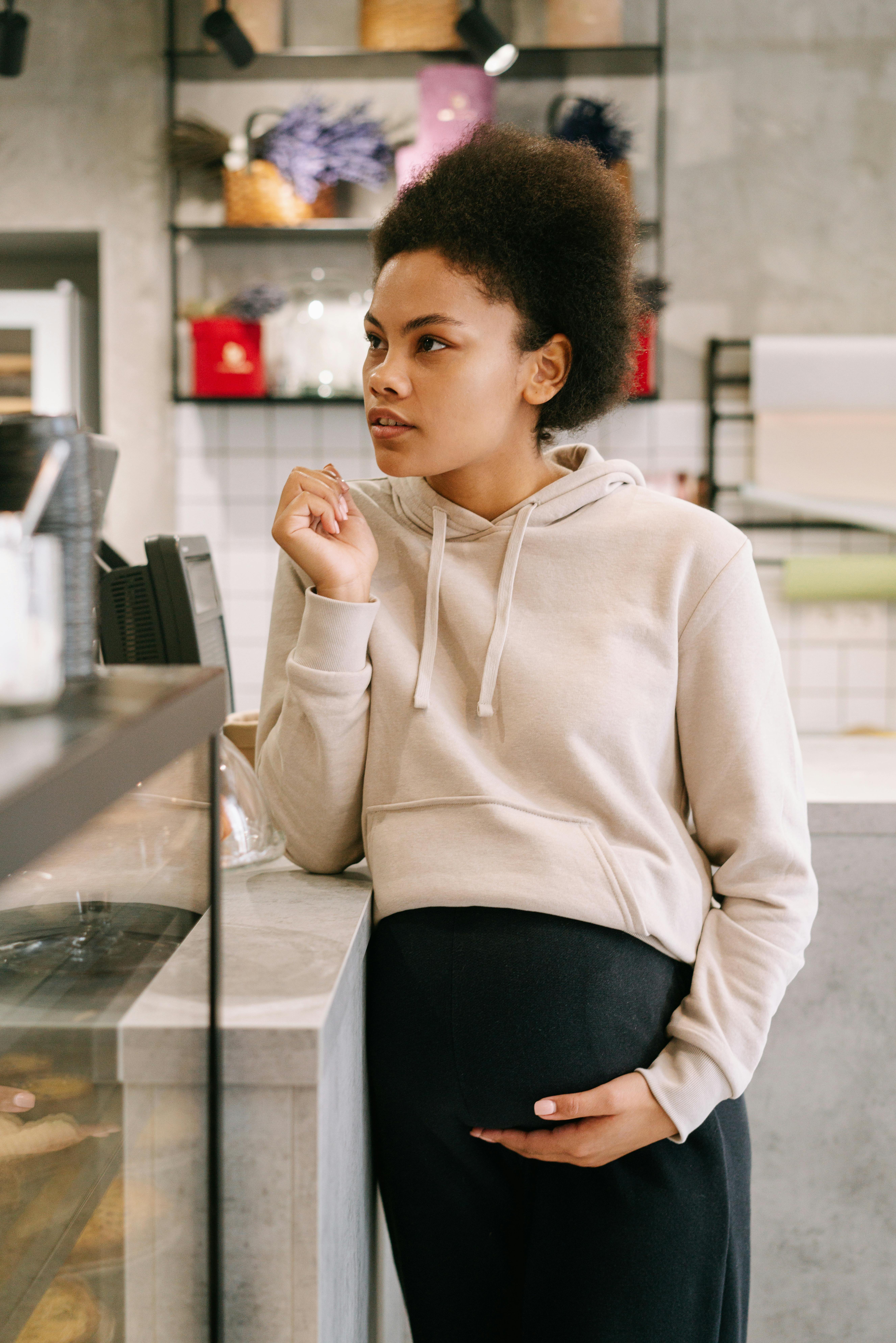 A pregnant woman stands indoors, leaning on a counter, wearing a hoodie and holding her belly.