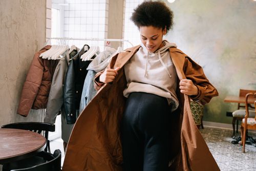 Free Pregnant woman in stylish coat adjusting outfit indoors near clothes rack. Stock Photo