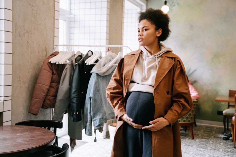 Pregnant woman wearing a stylish outfit in a modern indoor setting with coats in the background.