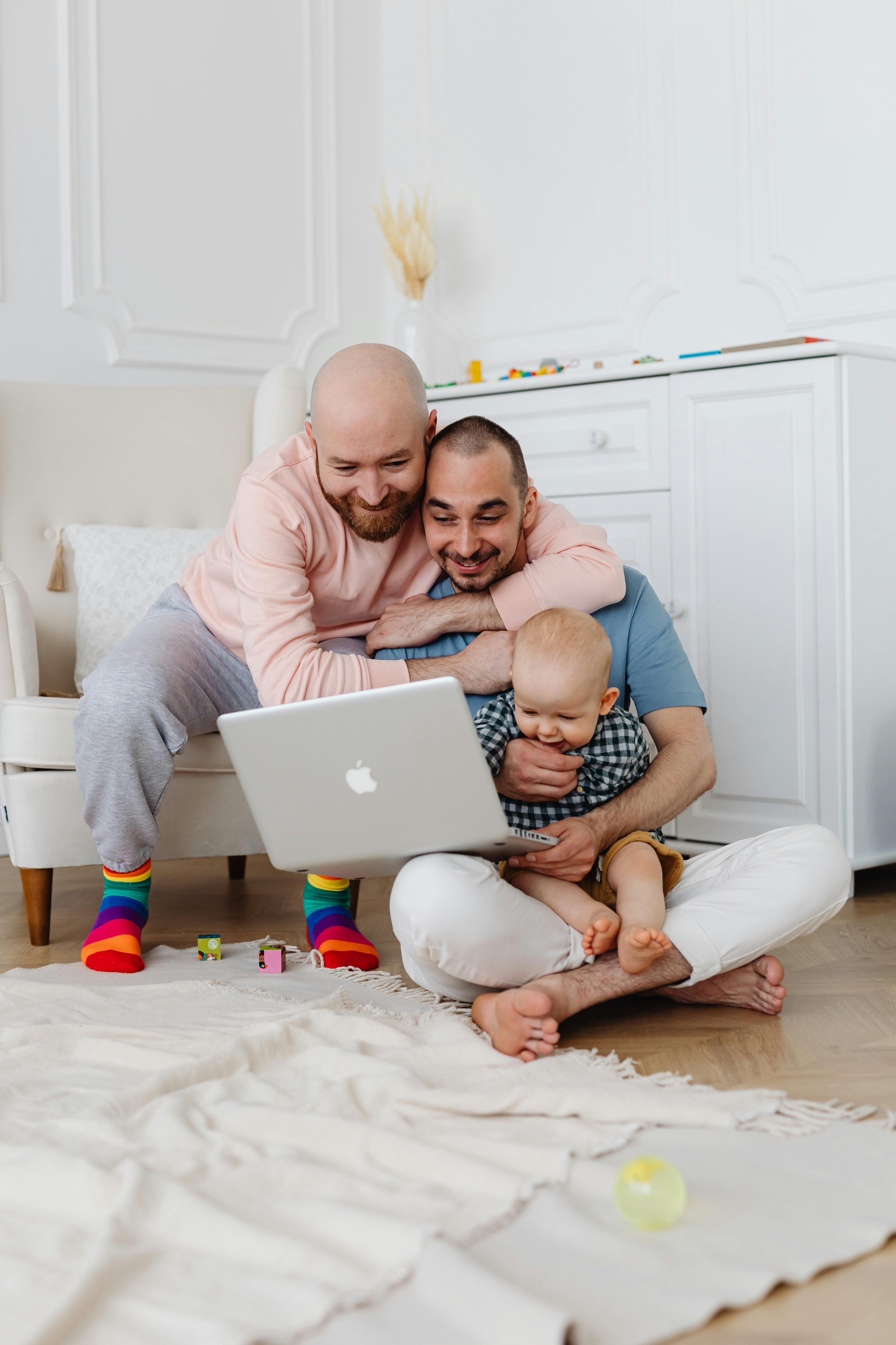 two men smiling while sitting on the floor with baby boy and looking at the laptop