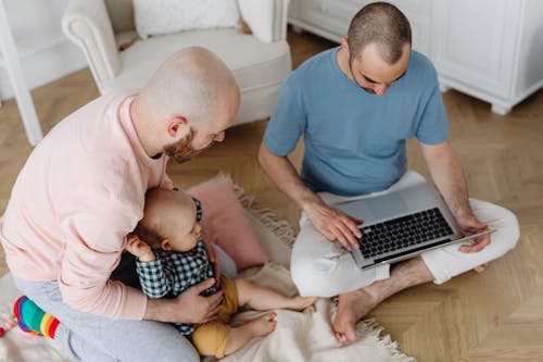 Free Man Holding Baby Boy Beside Man Sitting on the Floor Using Laptop Stock Photo