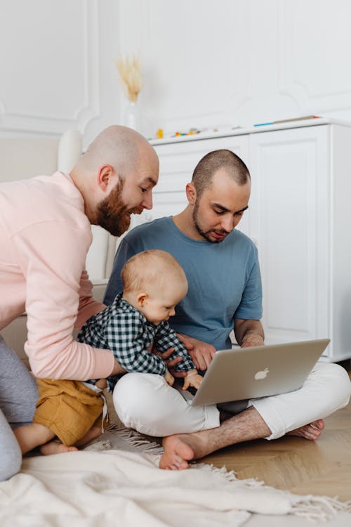 Free Man in Blue T-Shirt Using Laptop While Man in Pink Sweater Holding Baby Boy Beside Stock Photo