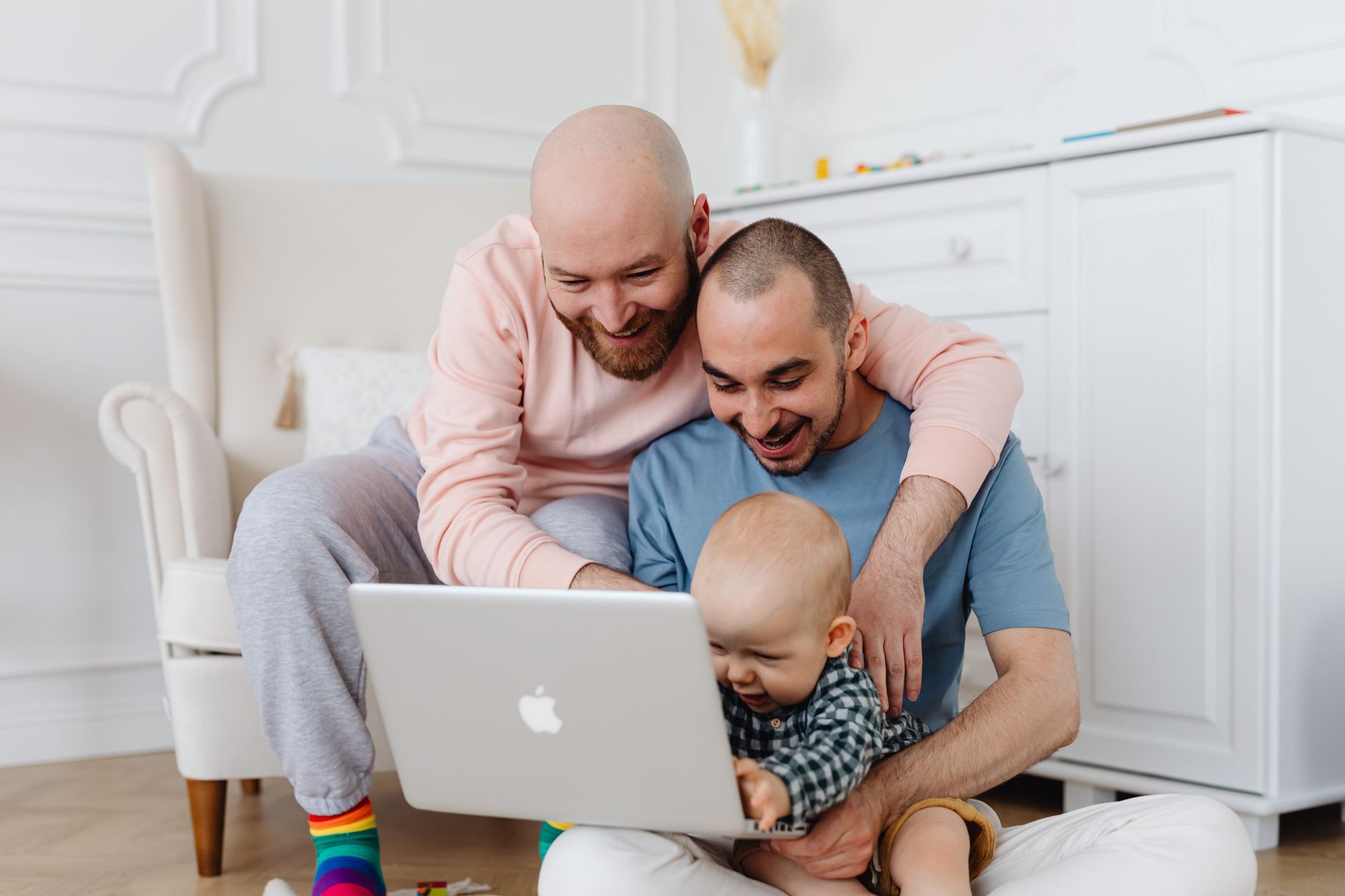 A joyful family scene indoors with two adults and a baby using a laptop, embracing and smiling.