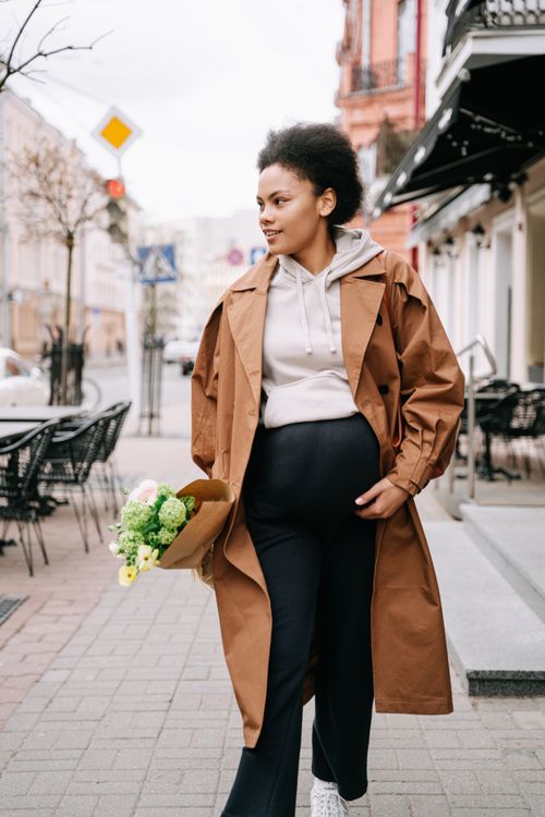 Free Pregnant woman with bouquet on city street wearing brown coat, capturing urban motherhood. Stock Photo