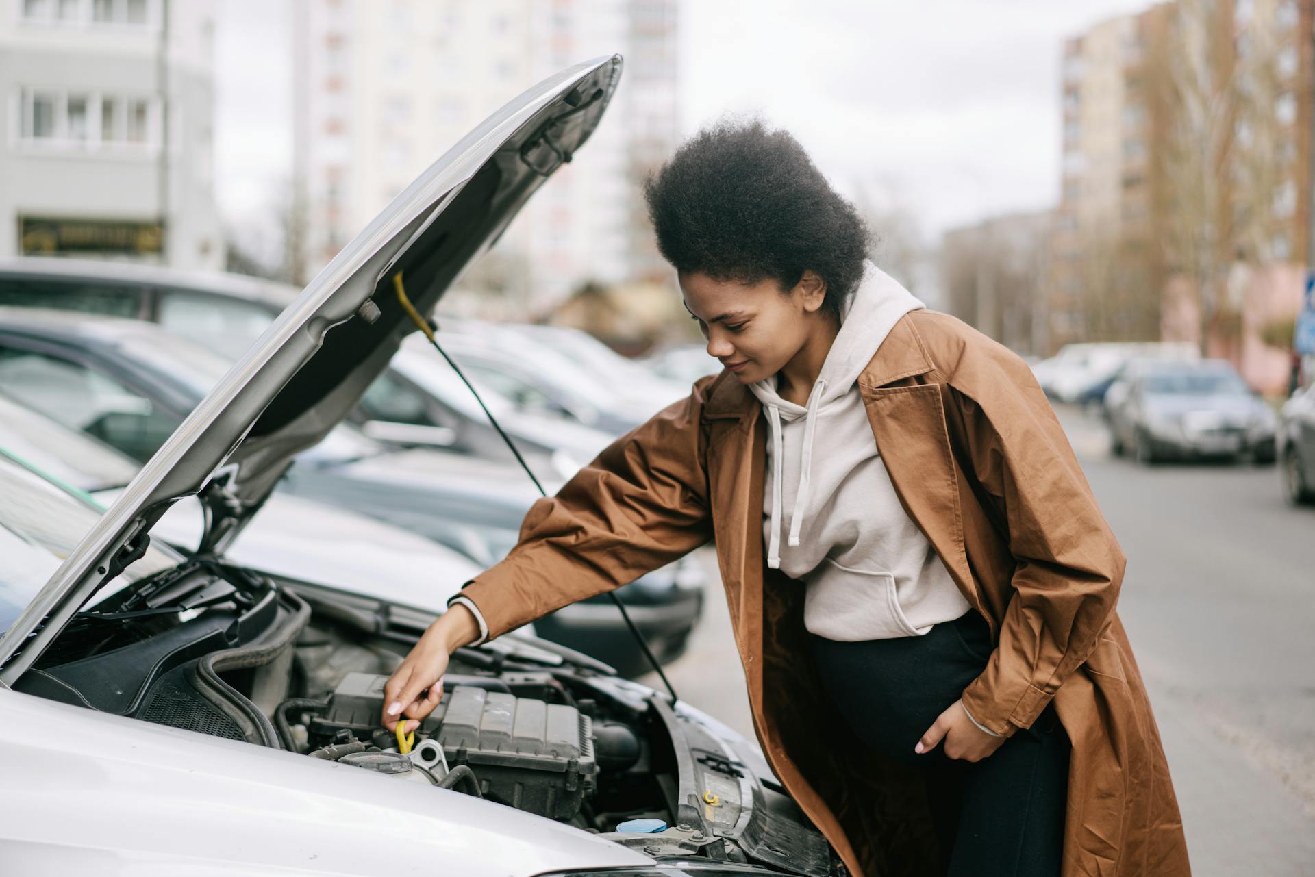 A pregnant woman inspects a car engine outdoors, showcasing independence and car maintenance skills.