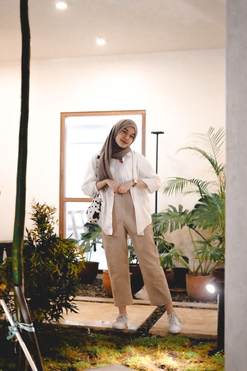 Free A stylish young woman in a hijab stands indoors surrounded by lush potted plants, exuding confidence and grace. Stock Photo
