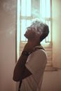 Side view of male in white t shirt and suspenders standing in light room with window and smoke at home