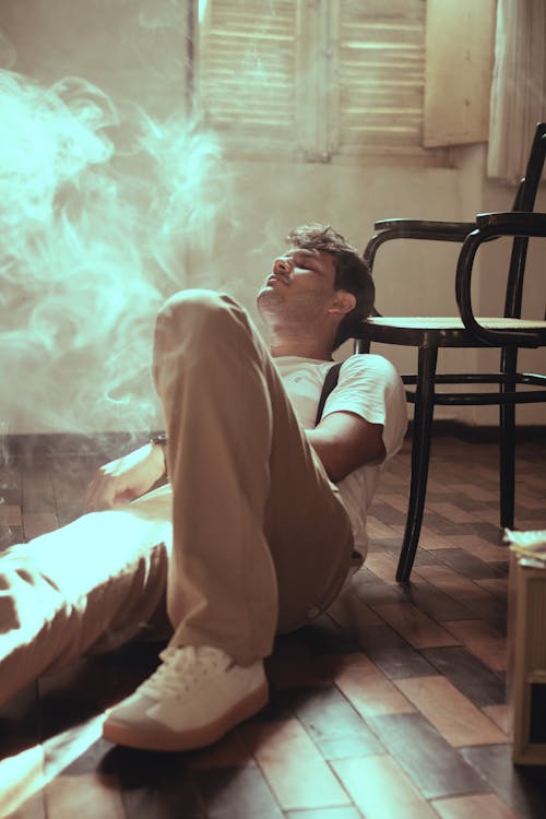 Calm male in stylish wear with suspenders lying on floor near chair in room with fume and shutters on window