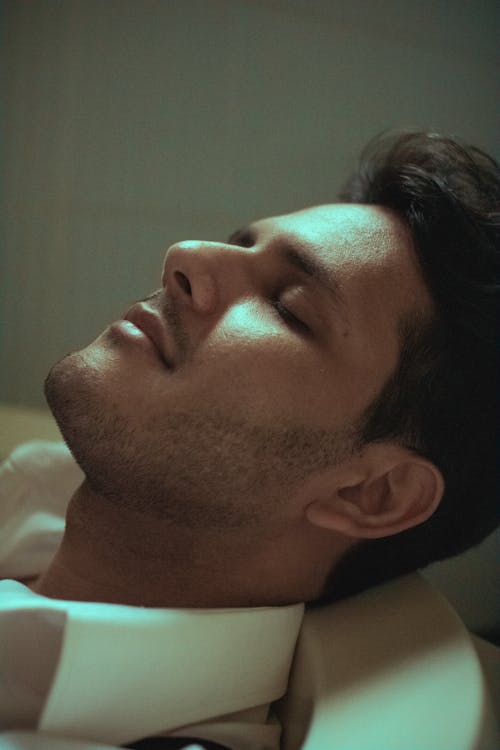 Side view of lonely calm male with closed eyes in white shirt lying in tub in with eyes closed in dim light