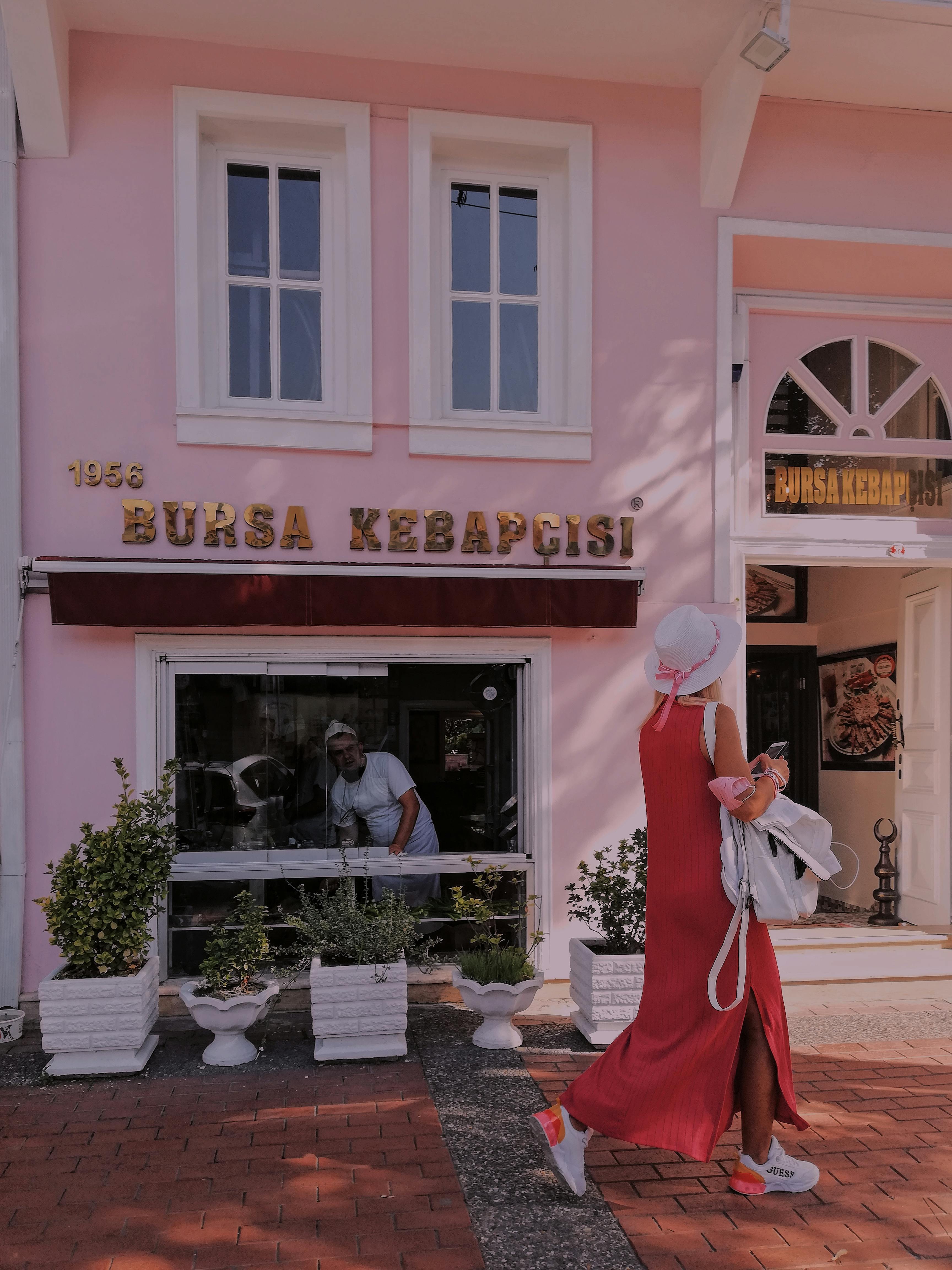 A stylish woman in a red dress walks by Bursa Kebabçısı café, featuring a pink wall and elegant urban setting.