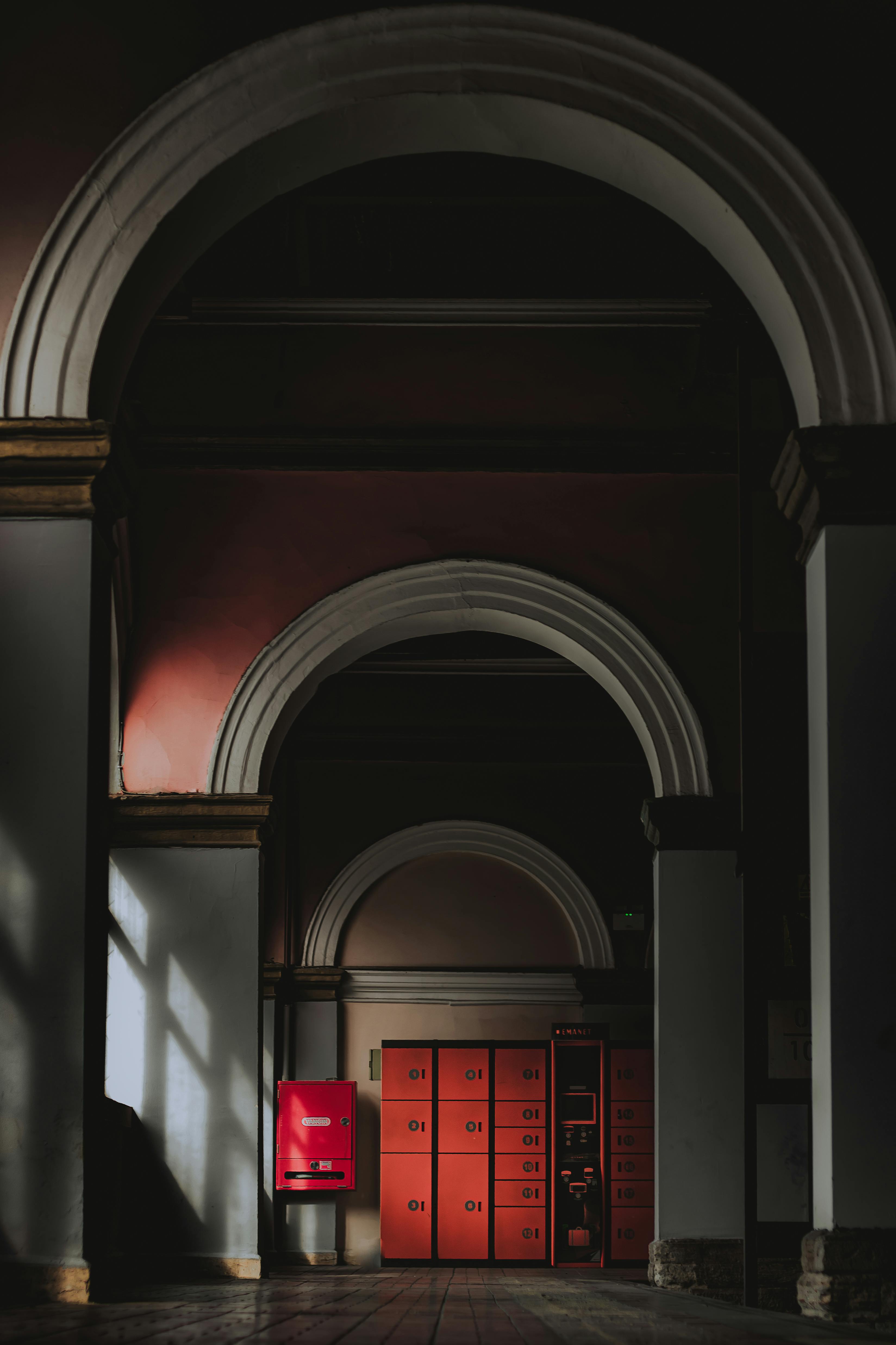 A mysterious indoor hallway featuring dramatic arches and red lockers, casting intriguing shadows.