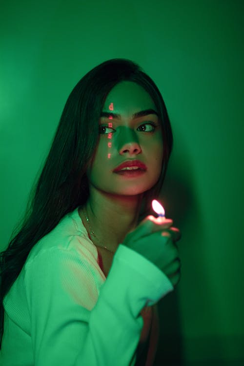 Dreamy female with dark hair and lighter in hand looking away while standing in dark room with glowing inscription on face